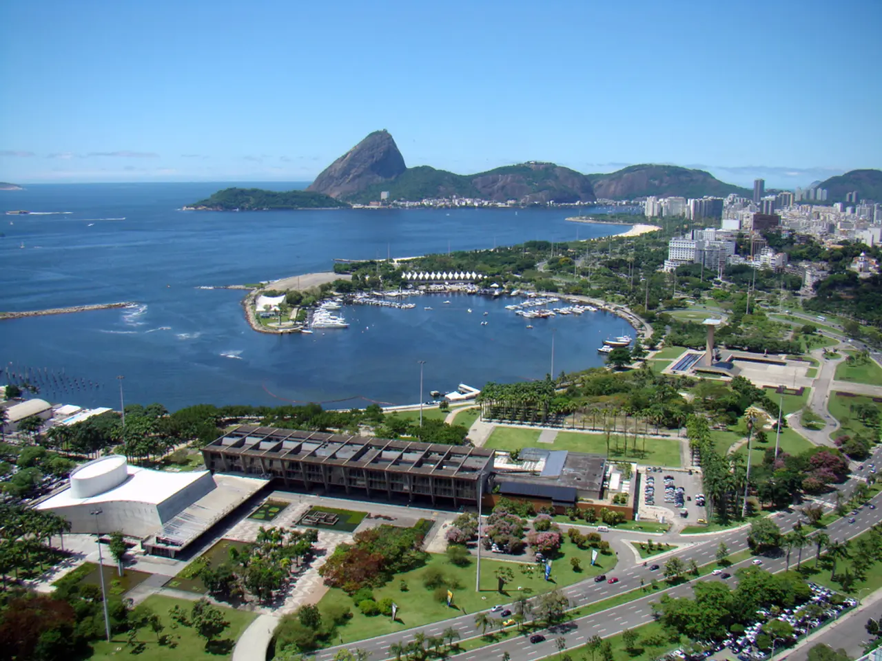 This image is an aerial view. In this image we can see many buildings, roads, trees, vehicles, sea,...