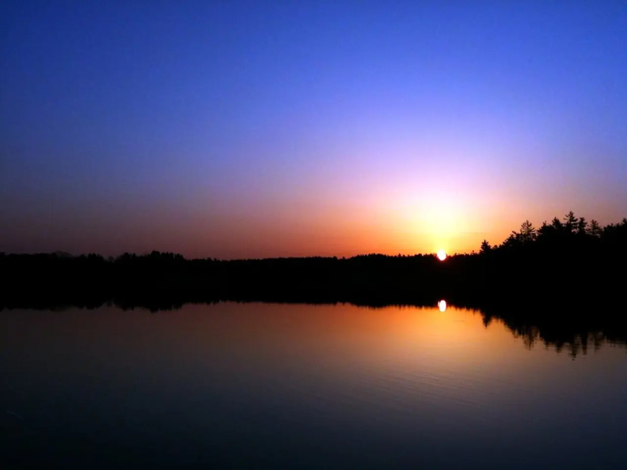 In the picture we can see water and far away from it, we can see some trees, sun and sky which is...