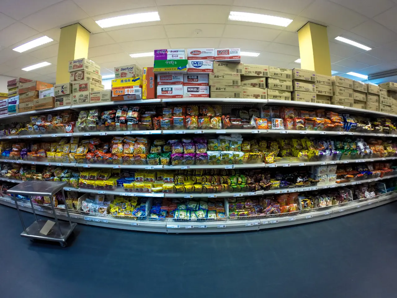 This is a mart. Here we can see food items and boxes on the racks on the floor. In the background...