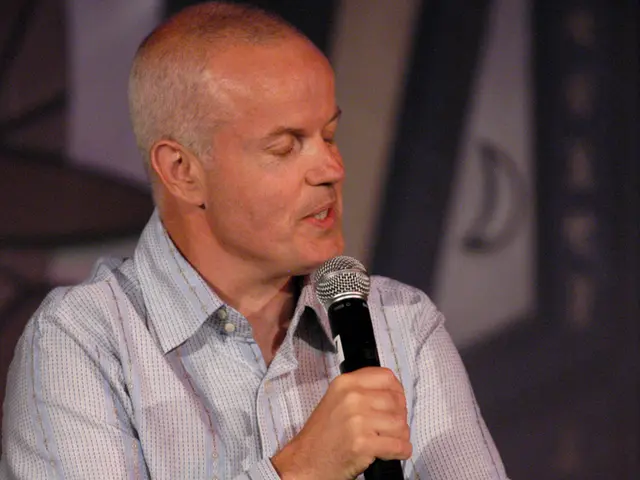 In the image there is a man sat holding a mic and talking on it.