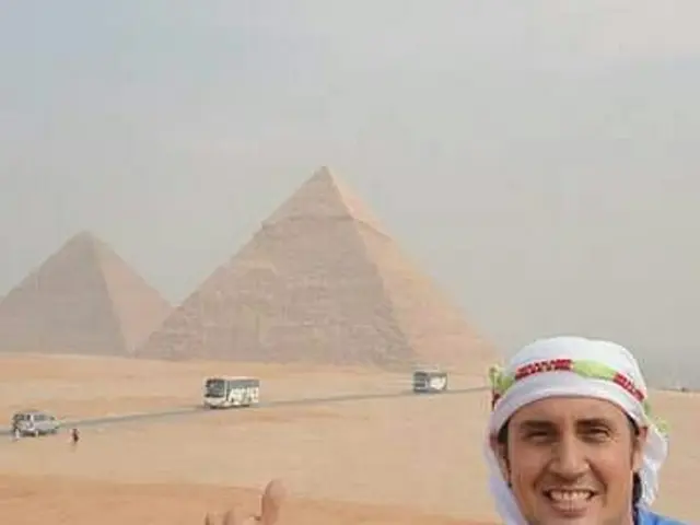 In this picture in the front there is a man smiling. In the background there are vehicles on the...