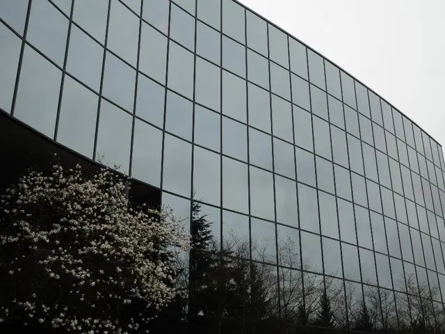 In the image there is a building with glass panels all over it with trees in front of it and above...