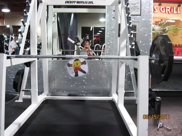 This image is clicked in a gym. In the front, there is a stand alone with dead weights and rod. At...