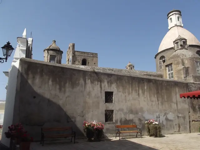 Here we can see a building,windows and at the wall there are benches and house plants. On the left...